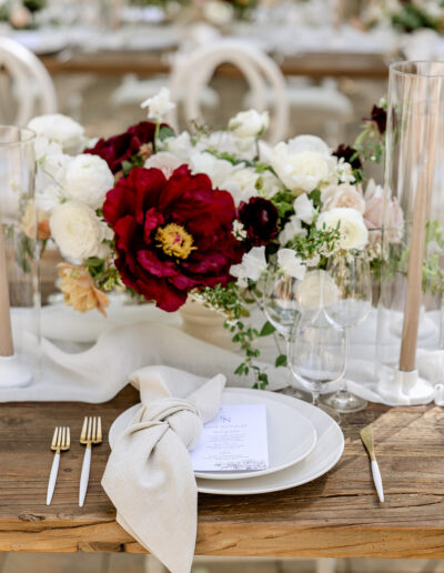 Elegant table setting with white plate, beige napkin, menu, gold flatware, and abundant floral centerpiece of red and white blooms, on a rustic wooden table.