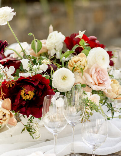 Elegant floral centerpiece with red, white, and pink flowers surrounded by clear wine glasses and candles on a white tablecloth.