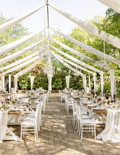 Outdoor wedding reception setup with long tables, white chairs, and flowing white drapes under a tented canopy. Tables are decorated with floral centerpieces, surrounded by trees.