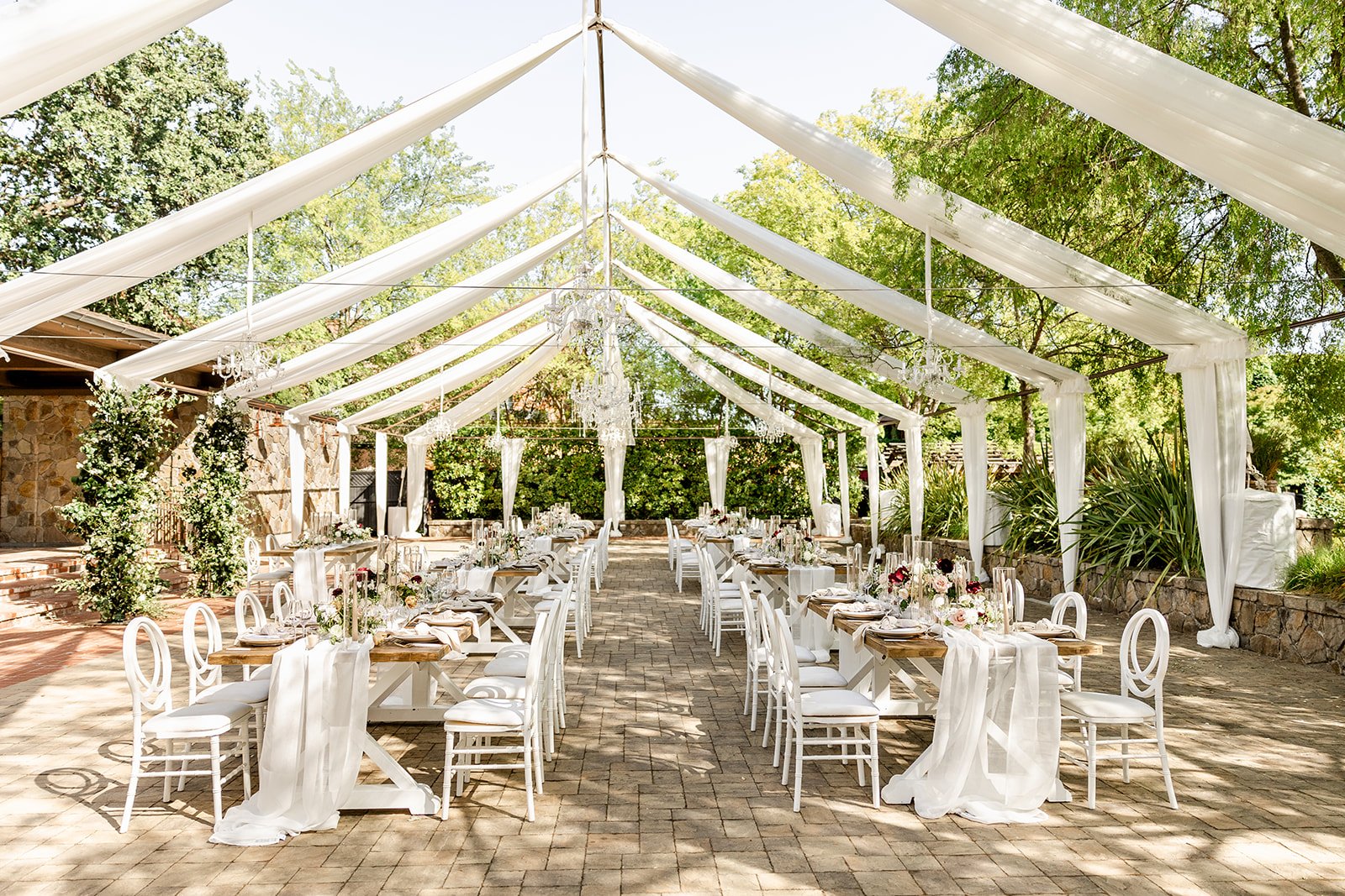 Outdoor wedding reception setup with long tables, white chairs, and flowing white drapes under a tented canopy. Tables are decorated with floral centerpieces, surrounded by trees.