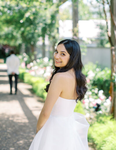 A woman in a white dress smiles while standing on a garden path, with flowers and trees in the background.