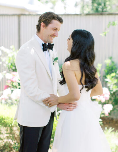 A bride and groom smile at each other in a garden setting, both wearing formal attire.