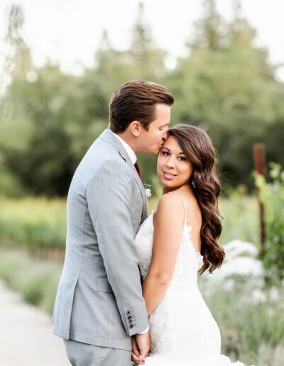 A couple embraces on a path in a garden. The man is wearing a gray suit, while the woman is in a white dress. They are surrounded by greenery and flowers.