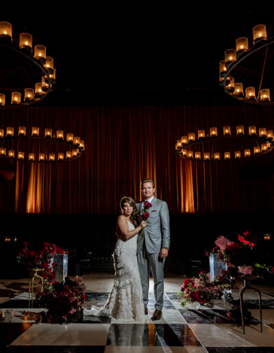 A bride and groom stand together on a dark, checkered floor with floral arrangements, under circular chandeliers with lit candles.