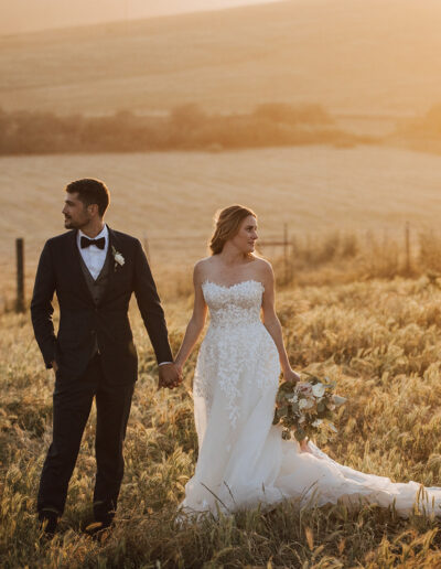 A bride and groom stand in a sunlit field, holding hands. The groom wears a dark suit and bow tie, while the bride wears a white gown and holds a bouquet.