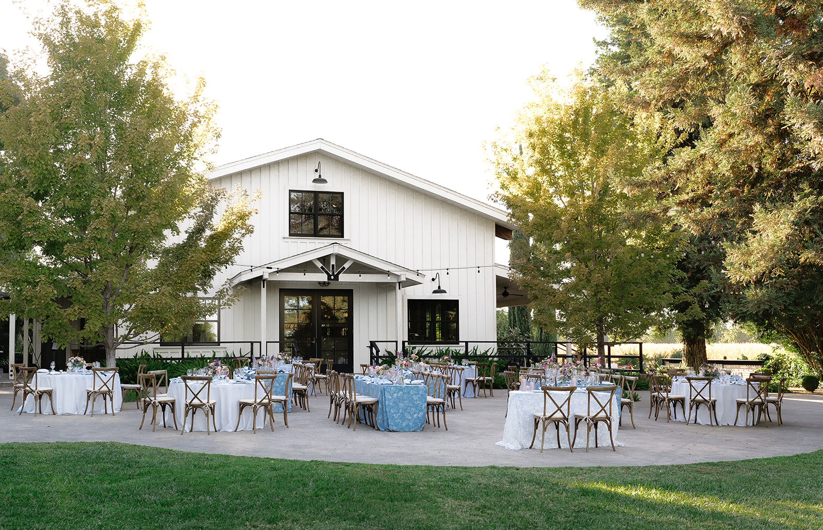 Round tables with chairs are set for an outdoor event in front of a white barn-style building surrounded by trees on a sunny day.