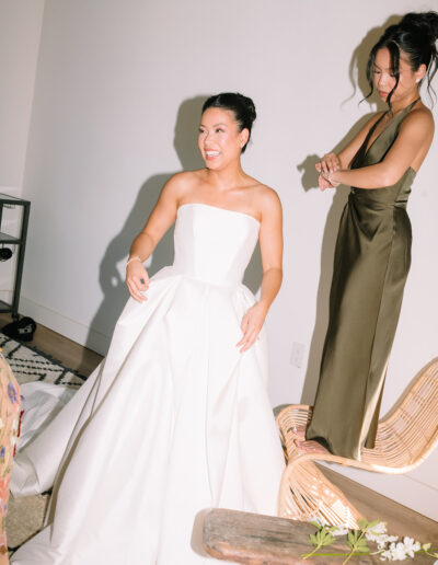A woman in a strapless white wedding dress smiles while another woman in a green satin dress stands nearby adjusting her bracelet.