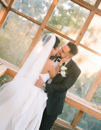 A bride and groom embrace and smile at each other in a sunlit wooden structure, with the bride wearing a long white veil and the groom in a black suit.