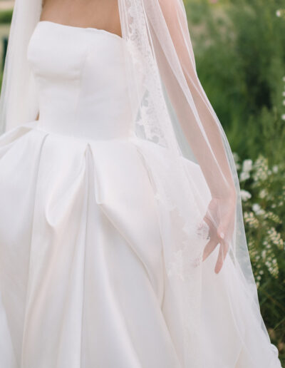 A bride in a strapless white wedding dress and lace-trimmed veil stands outdoors, surrounded by greenery and flowers.