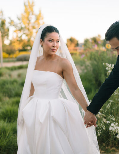 A bride in a white gown and a groom in a black suit hold hands outdoors, surrounded by greenery and flowers.