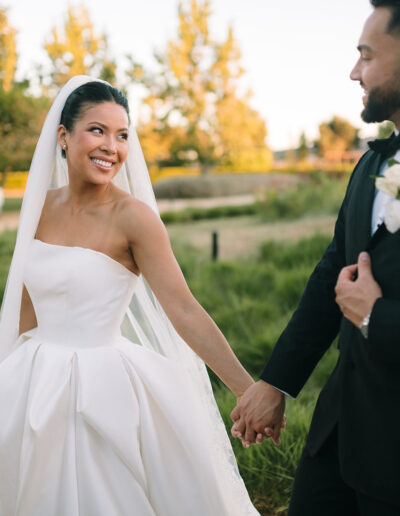 A bride in a white dress and veil holds hands with a groom in a black suit outdoors, both smiling amid green landscaping.
