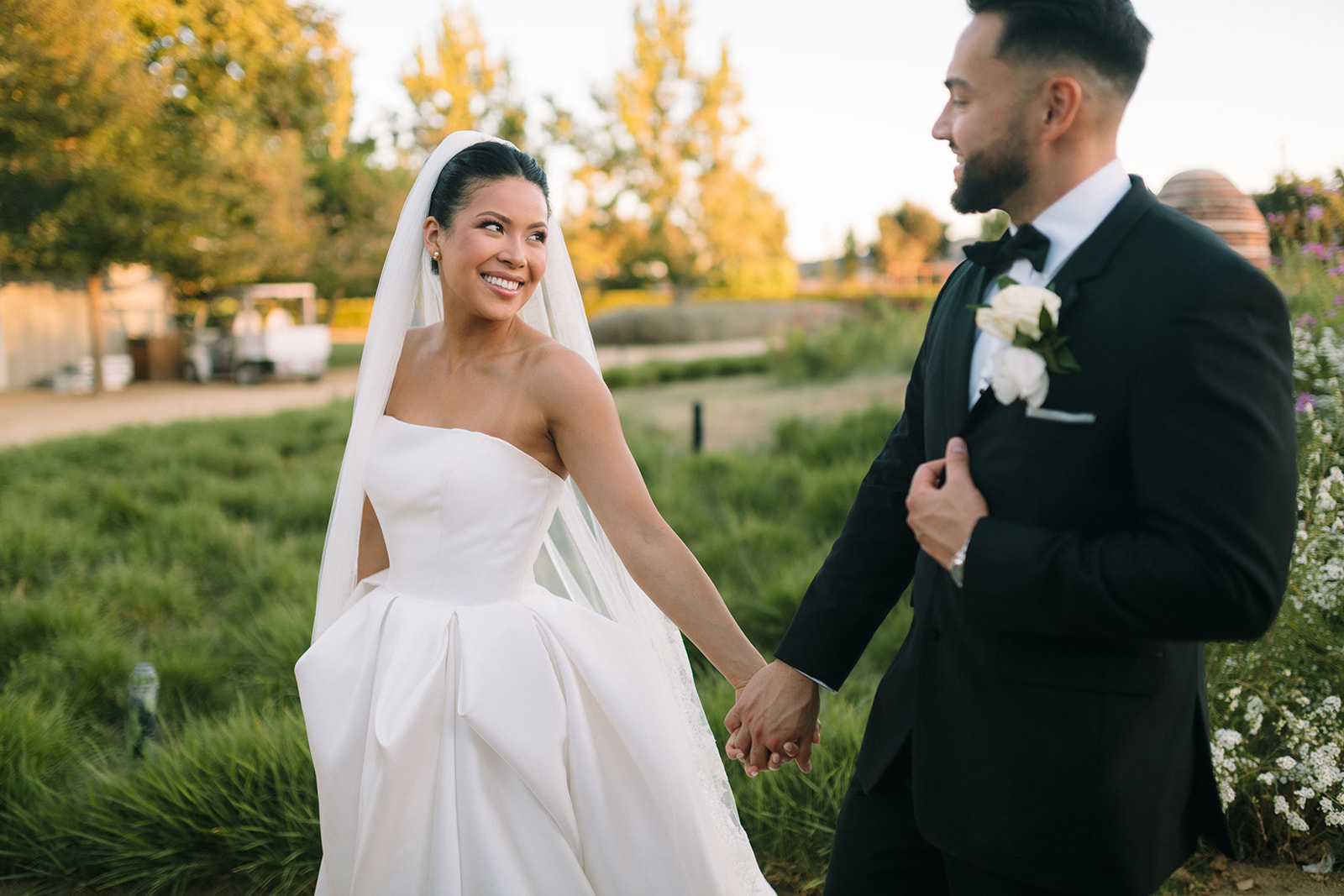 A bride in a white dress and veil holds hands with a groom in a black suit outdoors, both smiling amid green landscaping.