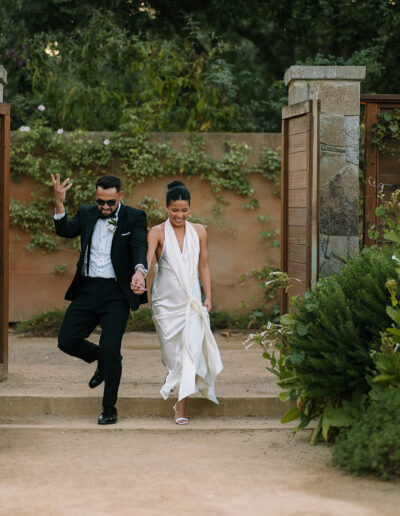 A couple in formal attire walks hand in hand down stone steps between open wooden gates, surrounded by lush greenery and flowers.
