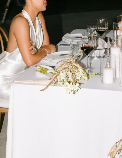 A woman in a white dress sits alone at a table set with candles, glassware, and a flower bouquet on a white tablecloth.