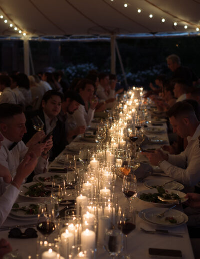 A long table under a tent is set for dinner with many people seated, eating and conversing, illuminated by numerous lit candles and string lights above.