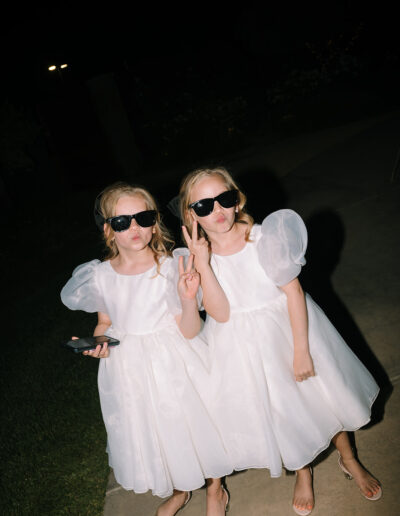 Two young girls in white dresses and sunglasses pose together at night; one holds a phone while the other makes a peace sign.