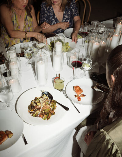 Three women sit at a candlelit dinner table set with salads, wine, cocktails, and water, engaged in conversation and serving food.