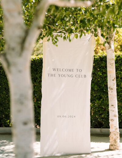 A cream-colored banner reading "WELCOME TO THE YOUNG CLUB" with the date "09.06.2024" is displayed outdoors between two trees.
