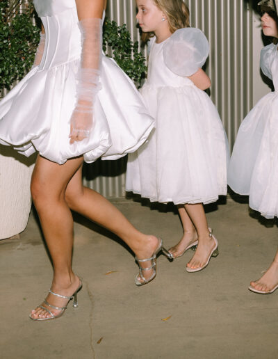 Three people in white dresses and heeled sandals walk outside on a concrete path, with two young girls following an adult. Potted plants and a tree are visible in the background.