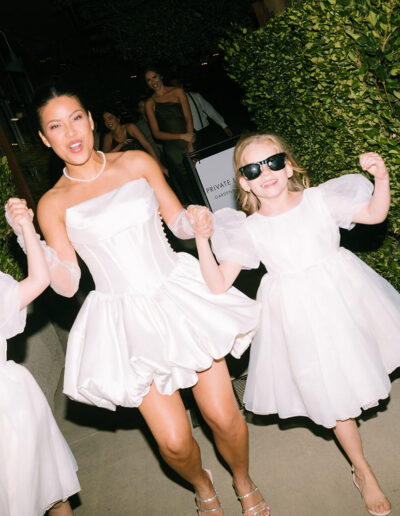 Three people wearing white dresses, including one adult and two children, walk together outdoors on a paved path lined with greenery at night.