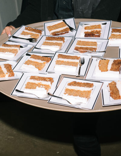 A person holds a large round tray with many square plates, each containing a slice of layered cake and a fork.