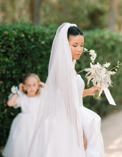 A bride in a white dress and veil holds a bouquet, while a young girl in a white dress stands in the background near green hedges.