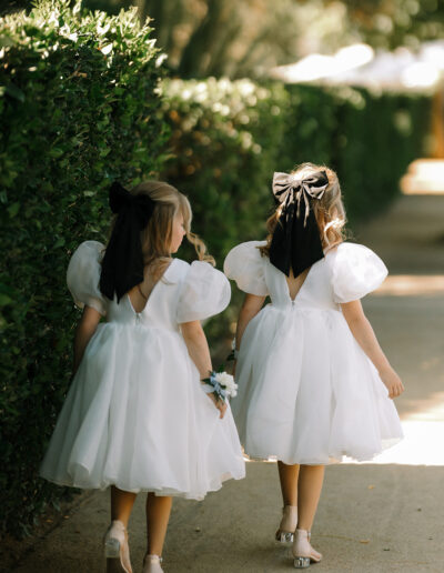 Two young girls in white dresses with puffed sleeves and large black hair bows walk side by side on a sunny path next to a green hedge.