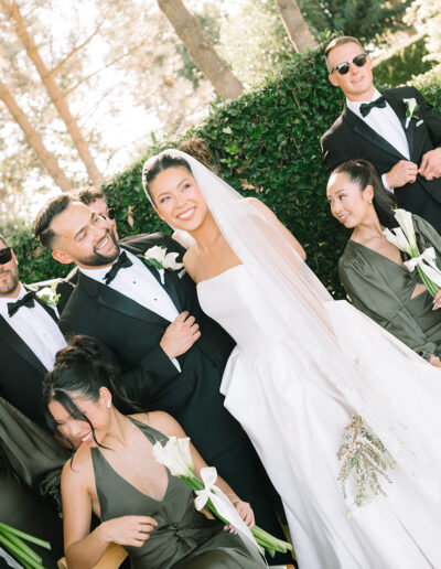 A bride and groom stand outdoors with their wedding party dressed in black and green, holding white flowers; a young girl with a parasol stands to the side.