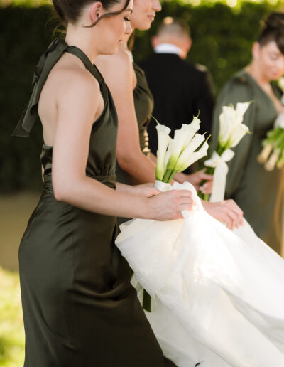 Two women in dark green dresses hold white calla lilies and assist with a white wedding dress outdoors, while another person stands in the background.