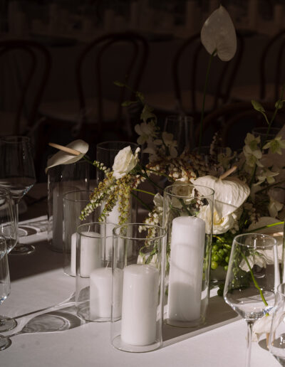 A close-up of a table set with white pillar candles in glass holders, white flowers, and glassware on a white tablecloth.