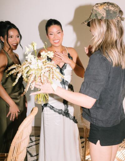 Three women indoors; one in a white dress with black trim holding flowers, another in a green dress, and a third in casual clothes with a camouflage hat.