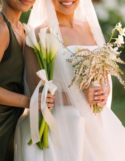 Two women pose together outdoors; one in a white wedding dress and veil holding a bouquet, the other in a dark dress holding calla lilies tied with ribbon.
