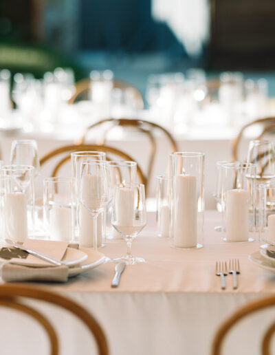 A formal table setting with white tablecloths, multiple clear glasses, white plates, napkins, cutlery, and white pillar candles, arranged for an event or reception.