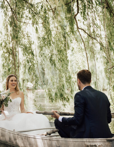 A bride in a white dress and a groom in a suit sit together in a canoe on a calm river, surrounded by hanging willow branches.