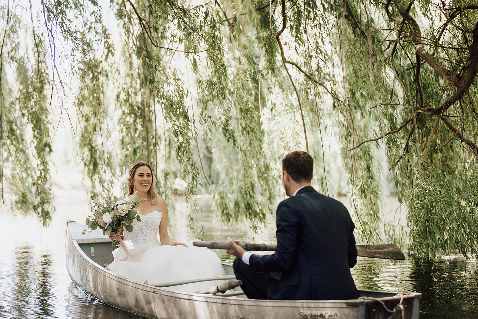A bride in a white dress and a groom in a suit sit together in a canoe on a calm river, surrounded by hanging willow branches.