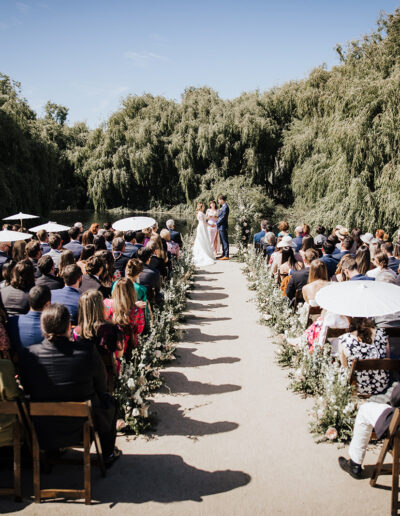An outdoor wedding ceremony with guests seated on either side of a flower-lined aisle, facing a couple standing with an officiant under clear skies and surrounded by trees.