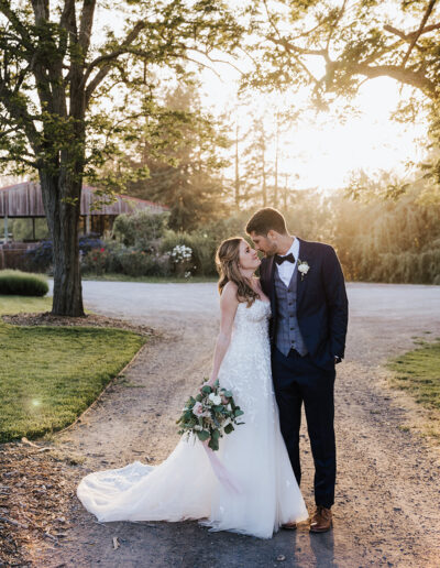 A bride and groom stand close together on a tree-lined path at sunset, with the bride holding a bouquet and the groom leaning in for a kiss.