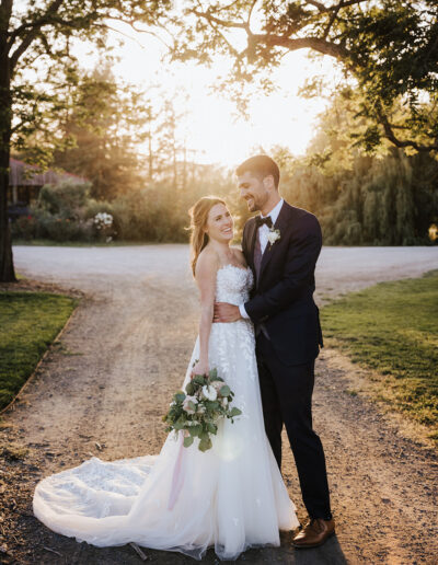 A bride and groom stand together outdoors at sunset, smiling, with the bride holding a bouquet and wearing a white dress, and the groom in a dark suit.