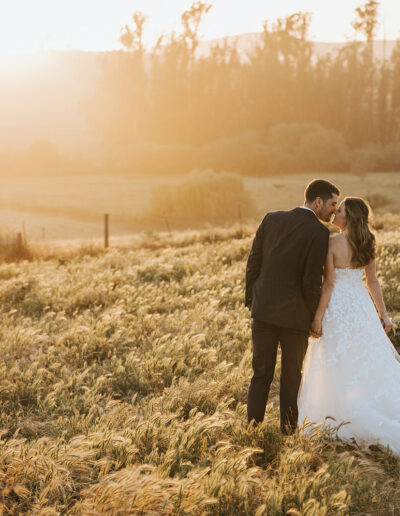 A bride and groom stand together in a grassy field at sunset, facing away from the camera, with trees and hills in the background.