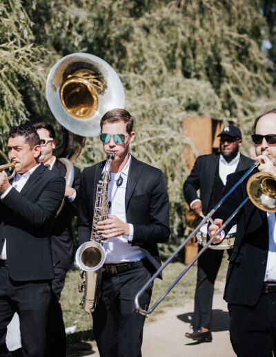 A brass band in black suits plays instruments, including trumpet, saxophone, trombone, and sousaphone, while marching outdoors along a tree-lined path.