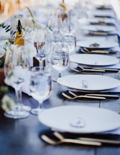 A long, elegant dining table set with white plates, glasses, cutlery, and floral centerpieces, ready for guests.