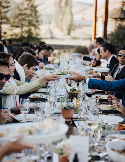 People seated at a long outdoor table share food and drinks during a formal gathering, with glassware and plated dishes visible.