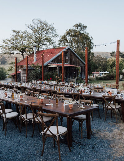 Long wooden tables with chairs are set up outdoors for an event near a rustic barn, with string lights overhead and countryside scenery in the background.