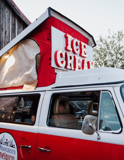 A red and white vintage van with a pop-up roof and large "ICE CREAM" sign is parked next to a rustic wooden building.