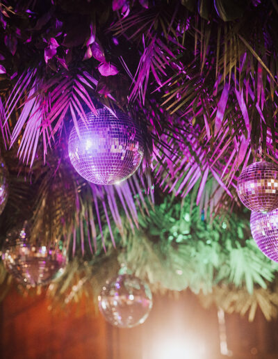 Disco balls hang from a ceiling decorated with green palm leaves and lit by purple and pink lights.