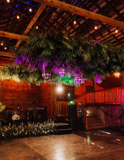 Large rustic barn interior with wooden beams, string lights, and greenery suspended from the ceiling; stage area and plants arranged along the floor.