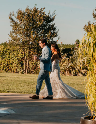A bride in a white dress and a groom in a blue suit walk side by side along a path near a pool, surrounded by greenery and trees.