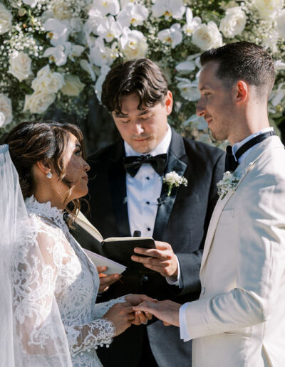 A bride and groom stand facing each other, holding hands, as an officiant reads from a book during an outdoor wedding ceremony in front of a floral arch.