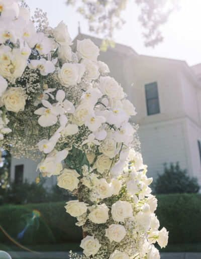 A floral arch made of white roses, orchids, and baby's breath stands outdoors in front of a white house on a sunny day.