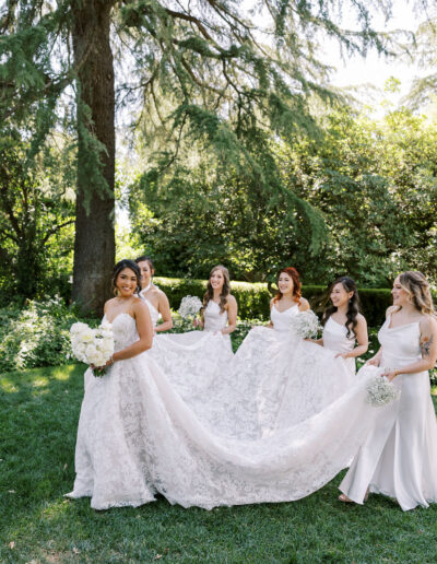 A bride in a lace gown holds a bouquet as four bridesmaids in white dresses help arrange her train outdoors under tall trees.